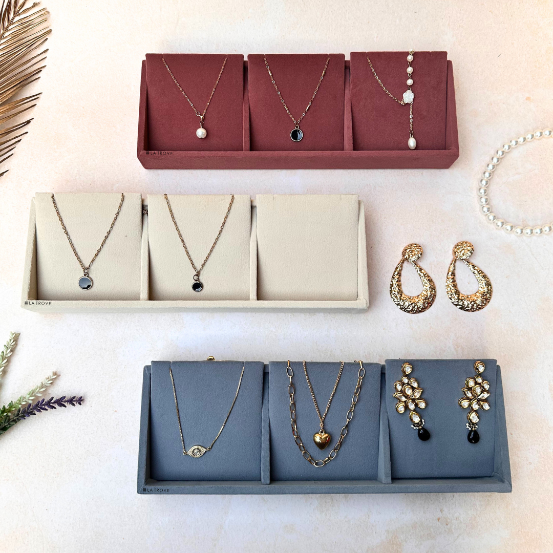 Three 3-slot velvet necklace organiser trays in dark pink, cream, and grey, each displaying gold chains and pendants.